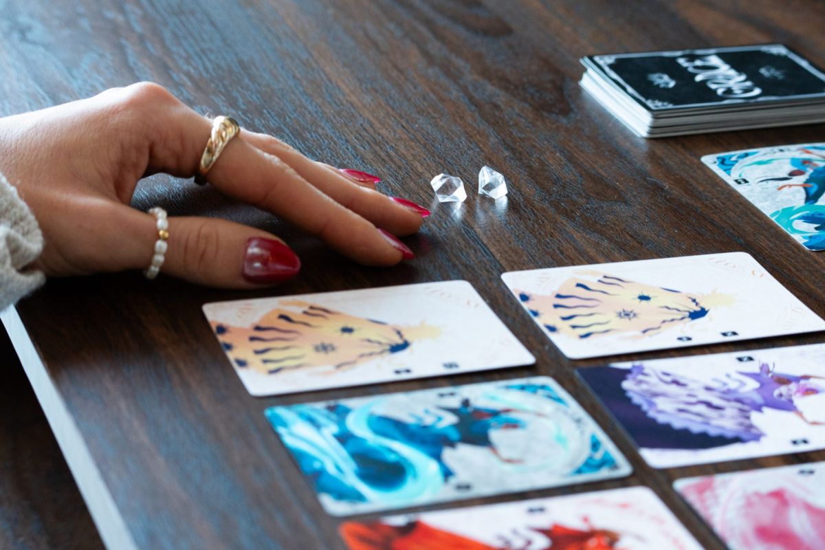 Une main posée sur une table en bois à côté de deux jetons de verre, devant des cartes illustrées étalées en grille.